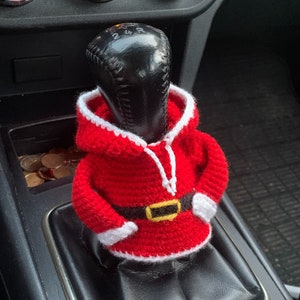 Puede incluir: Una funda de palanca de cambios de crochet roja en forma de Pap&aacute; Noel con ribete blanco y cintur&oacute;n negro. La funda est&aacute; en una palanca de cambios negra en un coche.