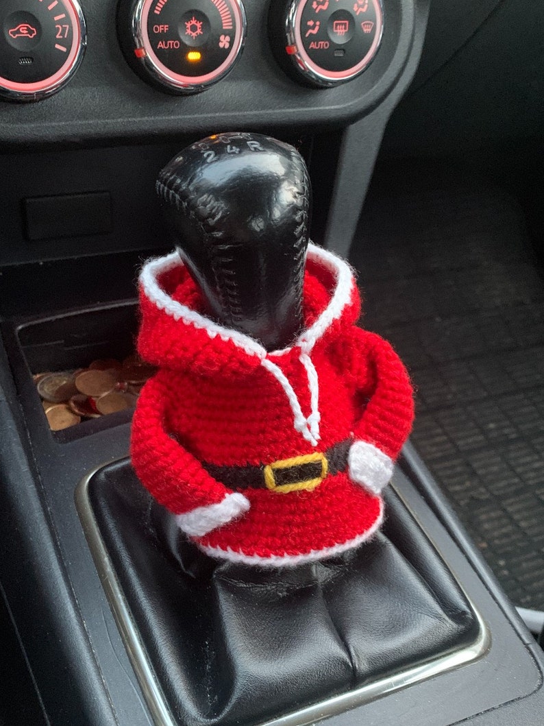Puede incluir: Una funda de palanca de cambios de crochet roja en forma de Pap&aacute; Noel con ribete blanco y cintur&oacute;n negro. La funda est&aacute; en una palanca de cambios negra en un coche.