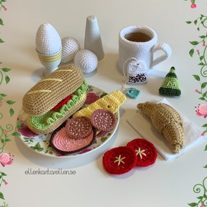 May include: A crocheted breakfast set with a sandwich, croissant, eggs, teacup, and tomato slices. The set is made of white, brown, green, red, and yellow yarn.