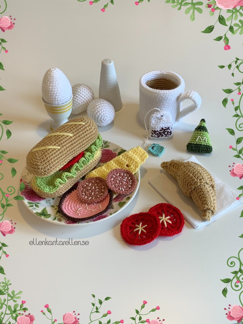 May include: A crocheted breakfast set with a sandwich, croissant, eggs, teacup, and tomato slices. The set is made of white, brown, green, red, and yellow yarn.
