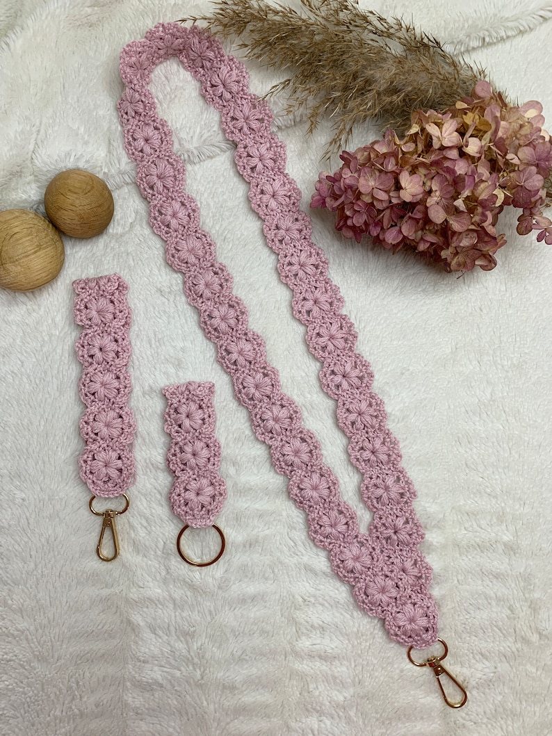 May include: A set of two pink crocheted keychains with gold key rings. The longer keychain is a lanyard style and the shorter keychain is a simple loop.
