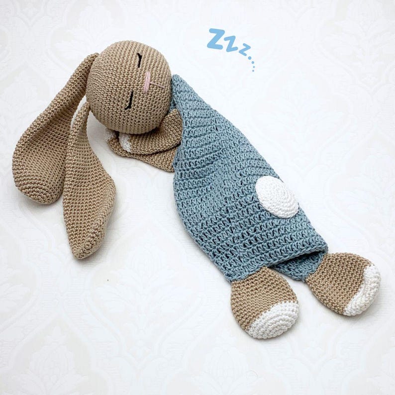 May include: A crocheted bunny doll in a sleeping pose. The bunny is primarily tan with long ears and closed eyes. It is covered by a blue blanket with a white patch. The word "Zzz..." is written above the bunny.
