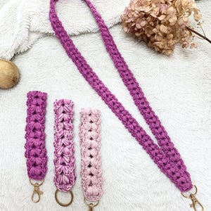 Pode incluir: Um cordão de crochê roxo feito à mão com um conjunto de chaveiros a condizer. O cordão e os chaveiros são feitos com um ponto texturizado e apresentam fechos giratórios dourados. O conjunto inclui um chaveiro roxo liso, um chaveiro roxo e branco e um chaveiro rosa claro.