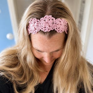 May include: A light pink crochet headband featuring a repeating floral pattern with textured, raised petals. The accessory is crafted from yarn and displays a series of interconnected, circular flower motifs that span across the front of the band.
