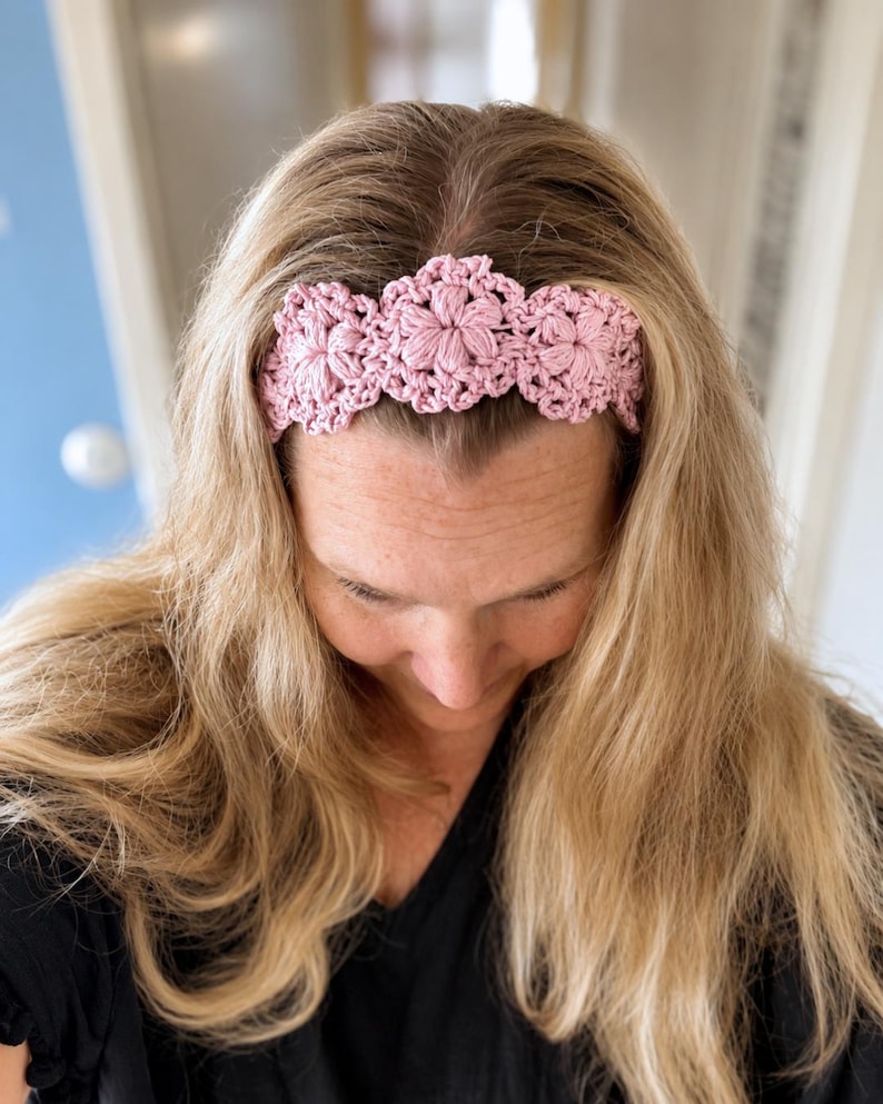 May include: A light pink crochet headband featuring a repeating floral pattern with textured, raised petals. The accessory is crafted from yarn and displays a series of interconnected, circular flower motifs that span across the front of the band.