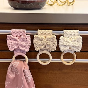 May include: Three handmade crochet towel holders in pink, beige, and cream, each featuring a bow design and a wooden ring. A pink patterned towel hangs from one holder. The holders are attached to a silver bar.