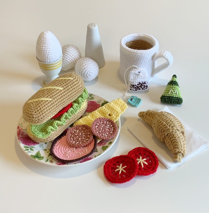 May include: A crocheted breakfast set with a sandwich, eggs, a cup of tea, and a croissant. The set is made of yarn and is a fun and whimsical way to decorate a kitchen or playroom.