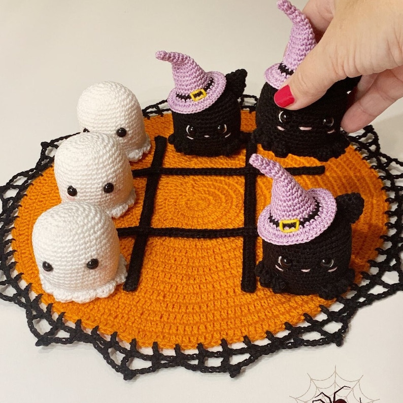 Puede incluir: Juego de tic-tac-toe de Halloween tejido a crochet. El juego presenta piezas de fantasmas blancos y piezas de gatos negros con sombreros de bruja morados. El tablero de juego naranja tiene un borde y una cuadr&iacute;cula negros.