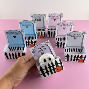 Puede incluir: Una colecci&oacute;n de cajas de regalo con tem&aacute;tica de Halloween con forma de l&aacute;pidas, cada una con un mensaje y dise&ntilde;o &uacute;nicos. Las cajas presentan un dise&ntilde;o de valla de piquetes negros, telara&ntilde;as e ilustraciones de calabazas. Una caja contiene un peque&ntilde;o fantasma blanco.