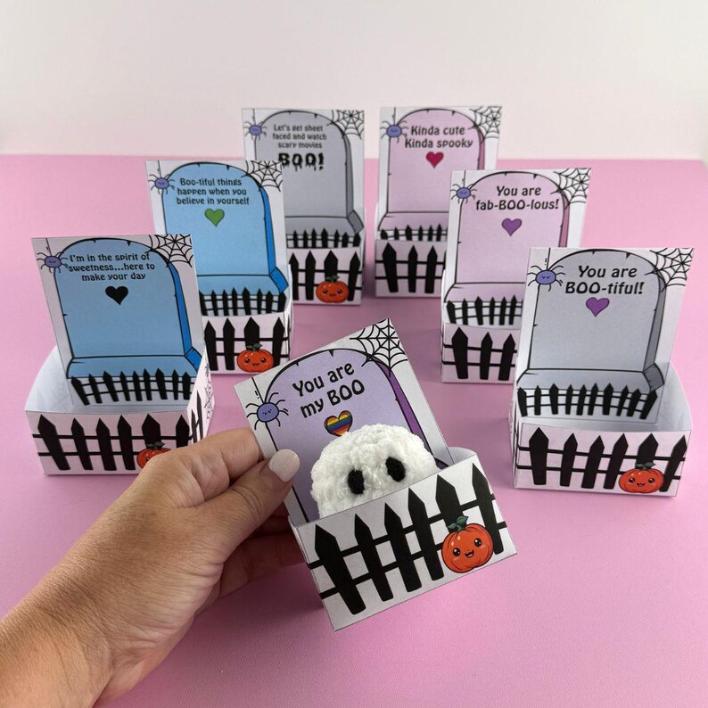 Puede incluir: Una colecci&oacute;n de cajas de regalo con tem&aacute;tica de Halloween con forma de l&aacute;pidas, cada una con un mensaje y dise&ntilde;o &uacute;nicos. Las cajas presentan un dise&ntilde;o de valla de piquetes negros, telara&ntilde;as e ilustraciones de calabazas. Una caja contiene un peque&ntilde;o fantasma blanco.