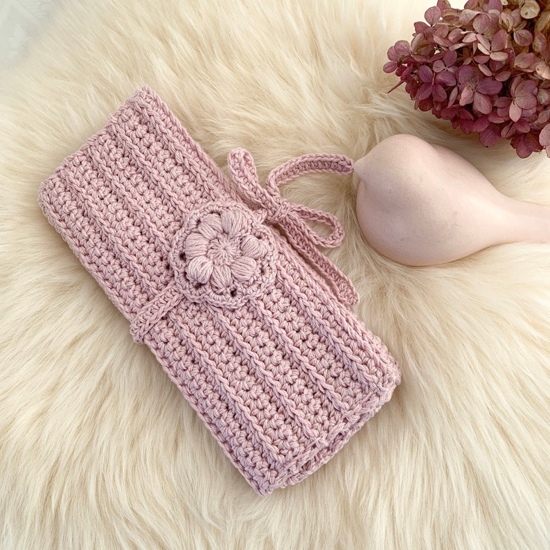 Puede incluir: Una bolsa de crochet rosa con un dise&ntilde;o de flores y un lazo. La bolsa est&aacute; sobre una superficie blanca y esponjosa.