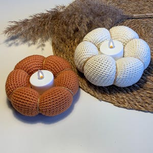 May include: Two crocheted candle holders, one in a light orange color and one in white, with a small LED tea light in each. The candle holders are shaped like a flower with multiple rounded petals.