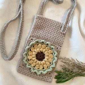 Könnte beinhalten: Beige gehäkelte Umhängetasche mit Sonnenblumenmuster. Die Sonnenblume hat eine braune Mitte, gelbe Blütenblätter und einen grünen Rand. Die Tasche hat einen langen, geflochtenen Riemen.