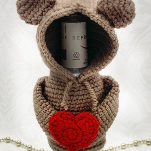 May include: A brown crocheted bear bottle cover with a red crocheted heart. The bear has a hood and large ears. The bottle cover is designed to fit a standard-sized bottle.