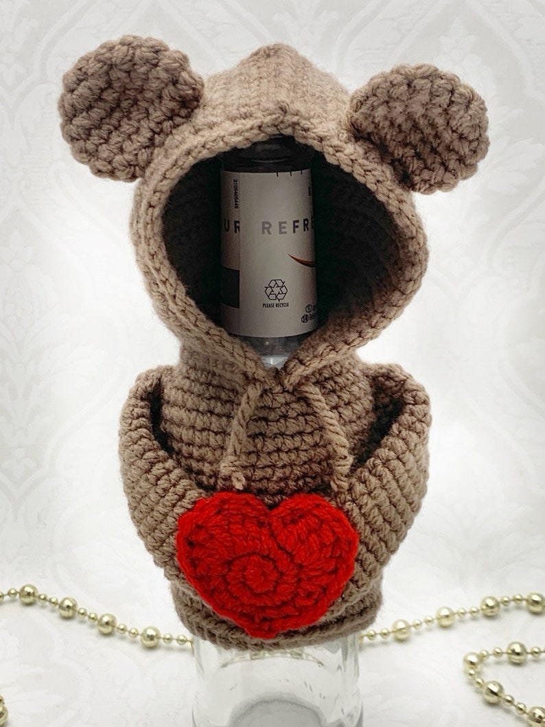 May include: A brown crocheted bear bottle cover with a red crocheted heart. The bear has a hood and large ears. The bottle cover is designed to fit a standard-sized bottle.