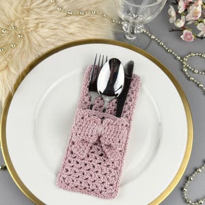 May include: A crocheted, mauve-colored cutlery holder with a bow detail, holding a fork, spoon, and knife. The holder is placed on a white plate with a gold rim, suggesting a formal dining setting. Decorative elements include a beaded garland and floral accents.