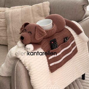 K&ouml;nnte beinhalten: Eine braune, geh&auml;kelte Hunde-Decke mit einer Tasche f&uuml;r eine Fernbedienung und einer wei&szlig;en Sch&uuml;ssel. Der Hund hat wei&szlig;e Pfoten und eine wei&szlig;e Schwanzspitze. Die Decke liegt auf einem wei&szlig;en, strukturierten &Uuml;berwurf.
