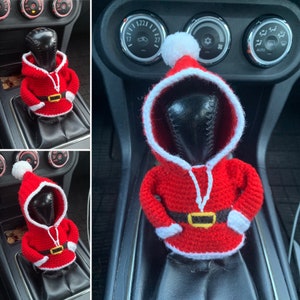 Pode incluir: Uma figura de Papai Noel de crochê em vermelho e branco, com um chapéu branco com pompom, é colocada na alavanca de câmbio de um carro.