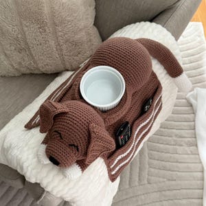 K&ouml;nnte beinhalten: Ein brauner, geh&auml;kelter, hundef&ouml;rmiger Couch-Caddy mit einer wei&szlig;en Sch&uuml;ssel, einer Fernbedienung und zwei Taschen. Der Hund hat wei&szlig;e Akzente und liegt auf einer wei&szlig;en Decke.