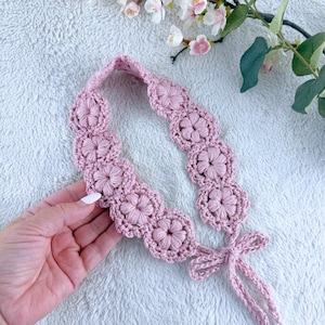 May include: A light pink crochet headband featuring a series of interconnected floral motifs. The accessory is constructed with a textured yarn pattern and includes two thin crocheted ties at one end that can be fastened into a bow.