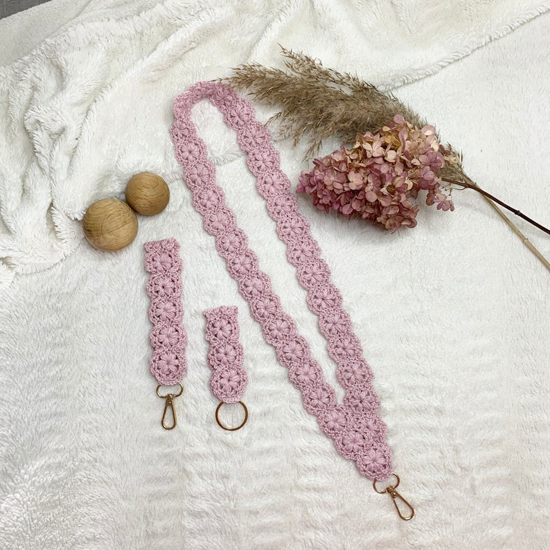 May include: Two pink crocheted keychains with gold key rings. The longer keychain is a strap with a flower pattern. The shorter keychain is a charm with a flower pattern. The keychains are on a white fluffy surface.