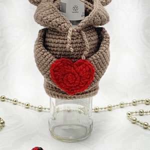 May include: A crocheted brown bear bottle cozy with a red heart in its paws. The bear is wearing a brown hooded sweater. A small red heart with black eyes is in the foreground.