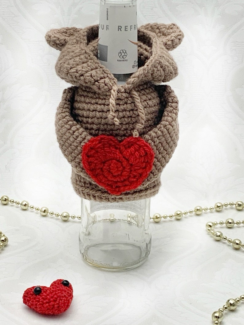 May include: A crocheted brown bear bottle cozy with a red heart in its paws. The bear is wearing a brown hooded sweater. A small red heart with black eyes is in the foreground.