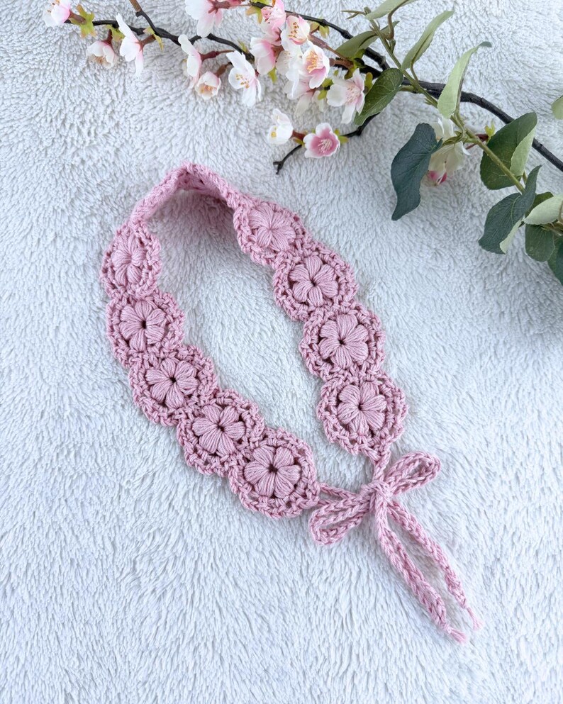 May include: A light pink crochet headband featuring a series of interconnected floral motifs with a textured, raised petal design. The headband is finished with two thin, matching crochet ties that form a bow at one end, crafted from soft yarn.