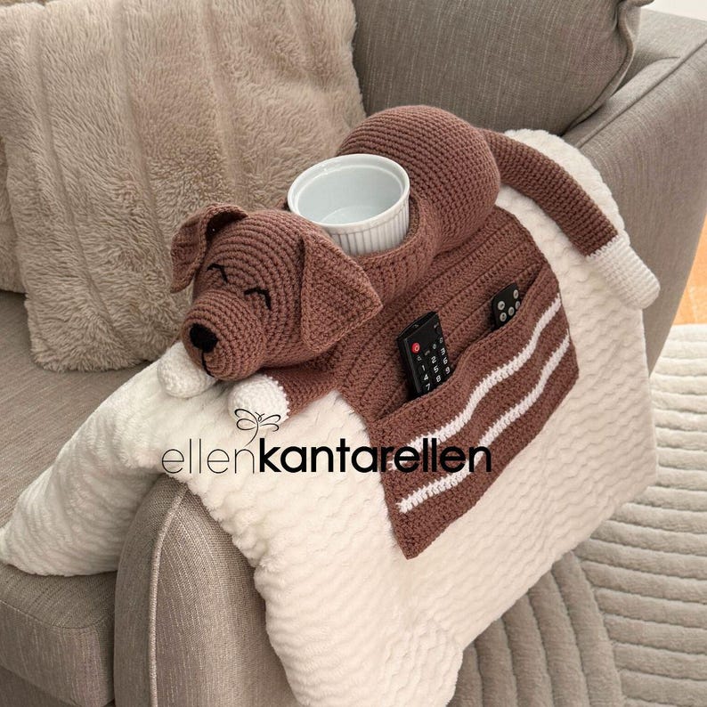 K&ouml;nnte beinhalten: Eine braun-wei&szlig;e geh&auml;kelte Hundedecke mit einer eingebauten Tasche und einer wei&szlig;en Sch&uuml;ssel. Die Decke hat einen braunen K&ouml;rper mit wei&szlig;en Akzenten und eine gestreifte Tasche. Eine Fernbedienung befindet sich in der Tasche. Die Decke liegt auf einem grauen Sofa.