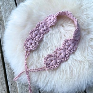 May include: A light pink crochet headband featuring a series of interconnected floral motifs with a textured, raised petal design. The accessory includes two thin, braided ties extending from the ends to allow for adjustable fastening around the head.