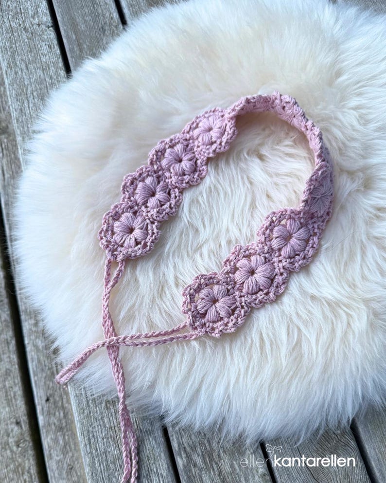 May include: A light pink crochet headband featuring a series of interconnected floral motifs with a textured, raised petal design. The accessory includes two thin, braided ties extending from the ends to allow for adjustable fastening around the head.