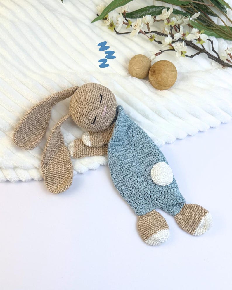 May include: A crocheted bunny toy in beige and light blue, lying on a white surface. The bunny has long ears, closed eyes, and a pink nose. It is covered with a light blue blanket with a white circle detail. Decorative wooden balls and flowers are in the background.
