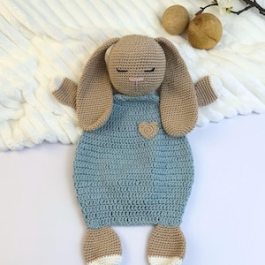 May include: A handmade crochet bunny toy in tan and light blue. The bunny has long ears, closed eyes, and a small heart detail on the front. The feet and tips of the ears are white. The toy is designed to be a baby comforter.