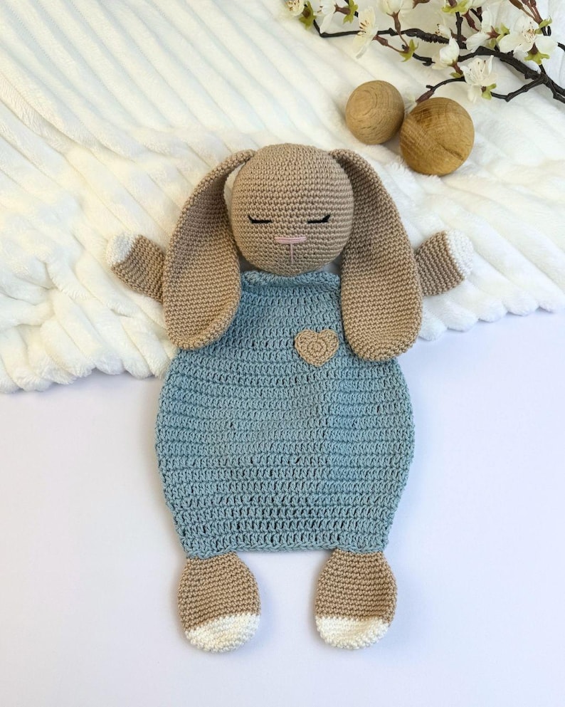 May include: A handmade crochet bunny toy in tan and light blue. The bunny has long ears, closed eyes, and a small heart detail on the front. The feet and tips of the ears are white. The toy is designed to be a baby comforter.