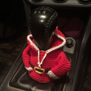 Puede incluir: Un disfraz de Pap&aacute; Noel de ganchillo para una palanca de cambios de coche. El disfraz es rojo con ribetes blancos y un cintur&oacute;n negro.