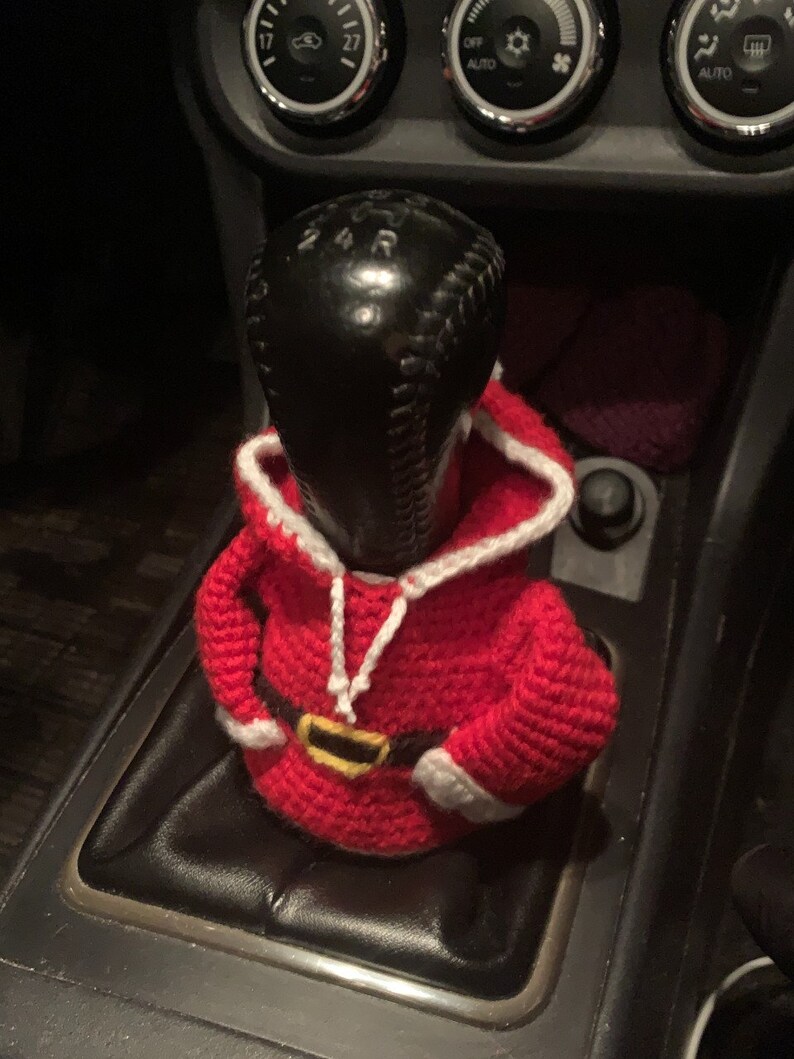 Puede incluir: Un disfraz de Pap&aacute; Noel de ganchillo para una palanca de cambios de coche. El disfraz es rojo con ribetes blancos y un cintur&oacute;n negro.