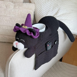 Puede incluir: Un organizador de brazo de sofá con forma de gato negro tejido a ganchillo con un sombrero de bruja. El gato tiene un hocico blanco y una nariz rosa. El organizador tiene bolsillos para un mando a distancia y otros objetos, y un portavasos.