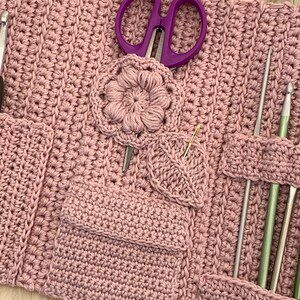 Puede incluir: Un estuche para agujas de crochet de color rosa con varios bolsillos y ranuras para guardar agujas de tejer, ganchos de crochet, tijeras y otras herramientas. El estuche tiene un dise&ntilde;o de flor y hoja de crochet.