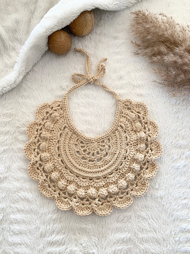 May include: A crocheted beige bib with a scalloped edge and a bow at the top.
