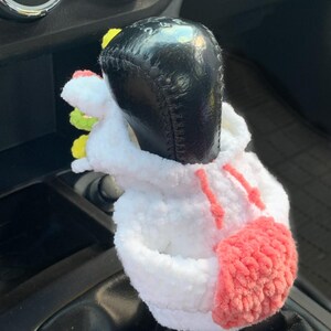 May include: A white crocheted unicorn gear shift cover with pink and yellow accents. The cover is on a black gear shift lever in a car.