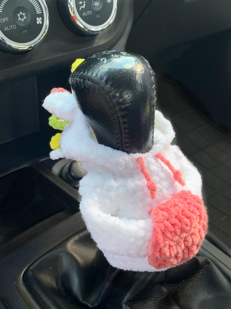 May include: A white crocheted unicorn gear shift cover with pink and yellow accents. The cover is on a black gear shift lever in a car.