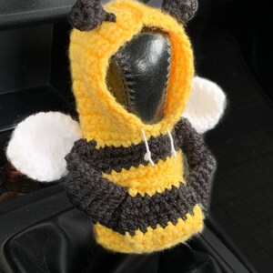 Peut inclure: Un couvre-levier de vitesse en crochet en forme d'abeille, jaune, noir et blanc. L'abeille a un corps ray&eacute; noir et jaune, des ailes blanches et une t&ecirc;te noire &agrave; capuche.
