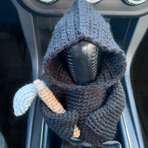 May include: A black crocheted figure of the Grim Reaper with a hooded robe and a small axe in its hand. The figure is attached to a car's gear shift lever.