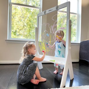 May include: Two children are drawing on a white double-sided easel with a clear dry-erase surface. The easel has a shelf for storing markers. The children are using colorful dry-erase markers.