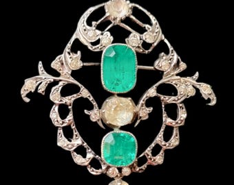 May include: A silver-toned brooch featuring an intricate openwork metal frame adorned with small clear stones. The piece is centered with two rectangular green stones and two round clear stones arranged in a vertical line.