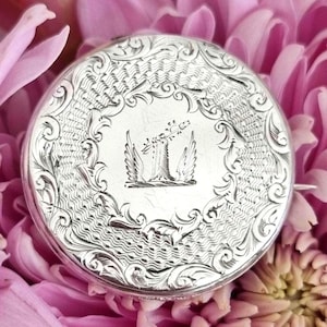 May include: A silver circular locket with an intricate engraved design. The locket features a central design of a bird with a crown above it.
