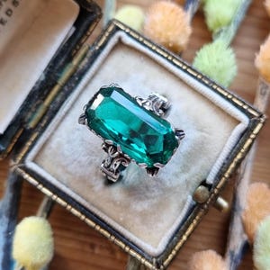 以下が含まれることがあります： 大きなエメラルドカットの緑色の宝石が付いた、ヴィンテージのシルバーリング。 リングは装飾的なシルバーバンドにセットされ、ベルベットの裏地が付いた小さなアンティークジュエリーボックスに収められています。 ボックスは閉じており、金色の留め金が付いています。