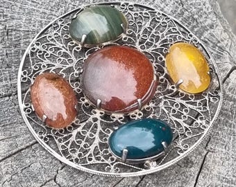 May include: A silver filigree brooch featuring five polished gemstones in shades of red, green, yellow, and blue. The oval gemstones are set in silver prongs within a circular filigree design. The brooch is displayed on a weathered wooden surface.
