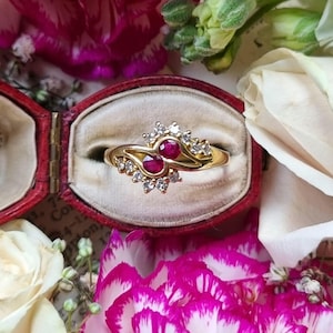 May include: A gold ring featuring two red gemstones and small clear stones, presented in a red velvet box. The ring is surrounded by pink and white flowers. The design of the ring has a swirling pattern.
