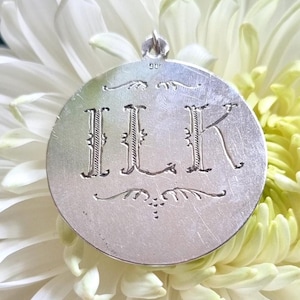 May include: Silver pendant with the initials "ILK" engraved in a script font. The pendant is circular and has a small loop at the top for attaching a chain.